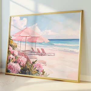 Vibrant framed painting of a dreamy beach scene leaning against a sunlit wall. The artwork depicts pink umbrellas and lounge chairs on pale sand, with lush pink flowers in the foreground and waves rolling in beneath a pastel sky.