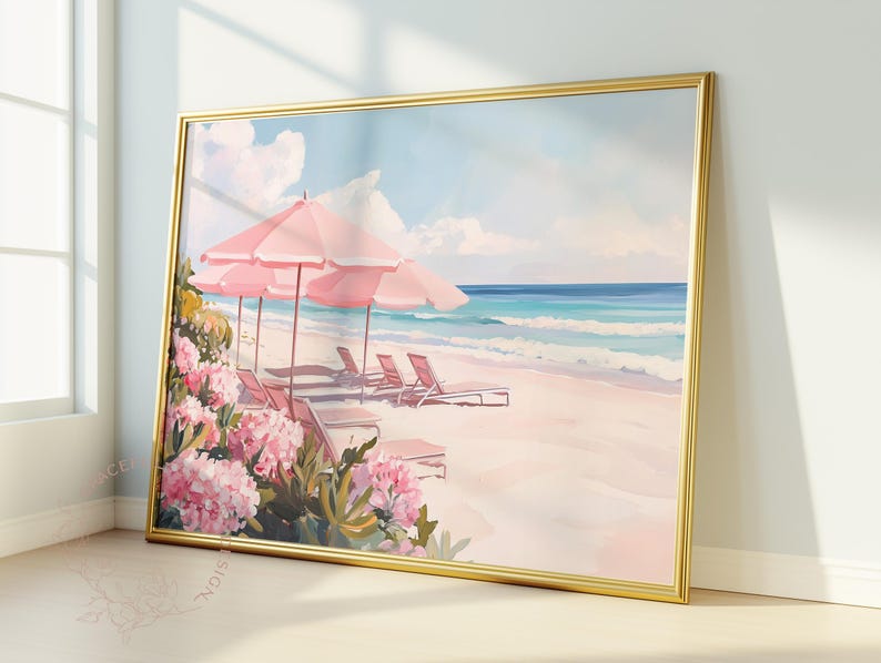 Vibrant framed painting of a dreamy beach scene leaning against a sunlit wall. The artwork depicts pink umbrellas and lounge chairs on pale sand, with lush pink flowers in the foreground and waves rolling in beneath a pastel sky.