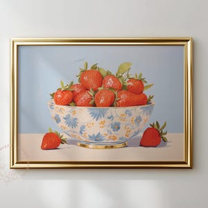 Framed painting of ripe strawberries piled in a white ceramic bowl decorated with blue and yellow flowers. A few strawberries rest beside the bowl on a flat surface. The background is soft blue, and the gold frame adds a refined touch to the still-life composition.