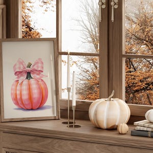 Puede incluir: Una impresi&oacute;n enmarcada de una calabaza rosa con un lazo de cuadros vichy se encuentra sobre una superficie de madera, junto a una calabaza blanca y una calabaza peque&ntilde;a. Dos velas blancas est&aacute;n en un candelabro dorado. La escena est&aacute; junto a una ventana.