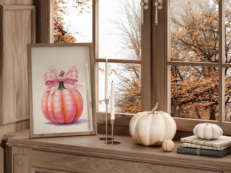 Puede incluir: Una impresi&oacute;n enmarcada de una calabaza rosa con un lazo de cuadros vichy se encuentra sobre una superficie de madera, junto a una calabaza blanca y una calabaza peque&ntilde;a. Dos velas blancas est&aacute;n en un candelabro dorado. La escena est&aacute; junto a una ventana.