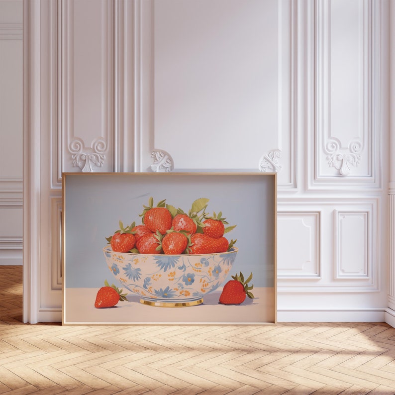 A framed painting of a bowl of strawberries, leaning against an ornate white wall with intricate molding. The bowl is decorated with blue and orange floral patterns, and a few strawberries sit outside the bowl on a light surface.