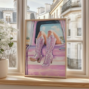 May include: A framed artwork depicts a person's legs and sparkly pink cowboy boots resting on a pink car. The boots have a high heel and are covered in glitter. The background shows a sunny sky and buildings.