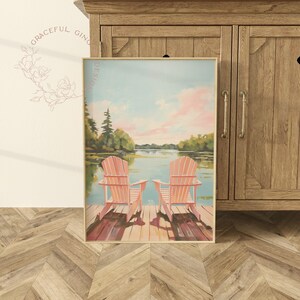 May include: A framed landscape painting featuring two pink Adirondack chairs on a wooden dock overlooking a lake. The sky is a soft blue with pink clouds, and trees line the distant shore. The artwork is displayed near a wooden cabinet.