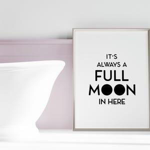 May include: A framed print with the text "IT'S ALWAYS A FULL MOON IN HERE" in black. The print is in a bathroom setting, next to a white bathtub and silver faucet.