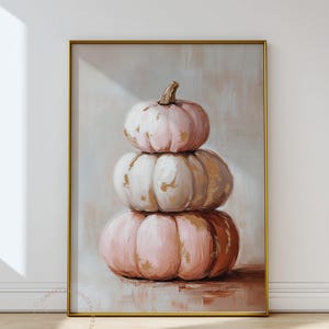 May include: A framed artwork depicting a stack of three pumpkins in pink, white, and brown hues. The pumpkins have a textured, painted look with gold highlights. The artwork is set against a neutral background.