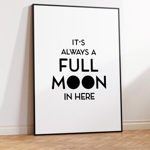 May include: A framed white poster with black text that reads "IT'S ALWAYS A FULL MOON IN HERE." The poster has a black frame and is leaning against a white wall on a wooden floor.