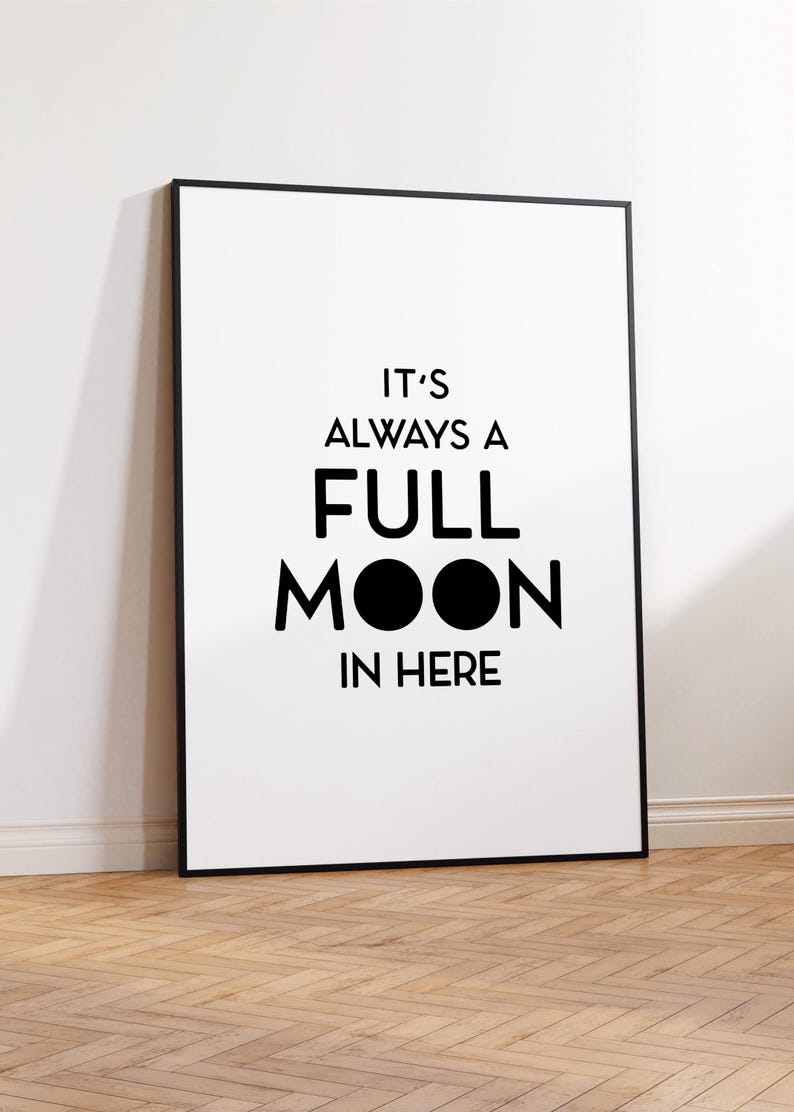 May include: A framed white poster with black text that reads "IT'S ALWAYS A FULL MOON IN HERE." The poster has a black frame and is leaning against a white wall on a wooden floor.