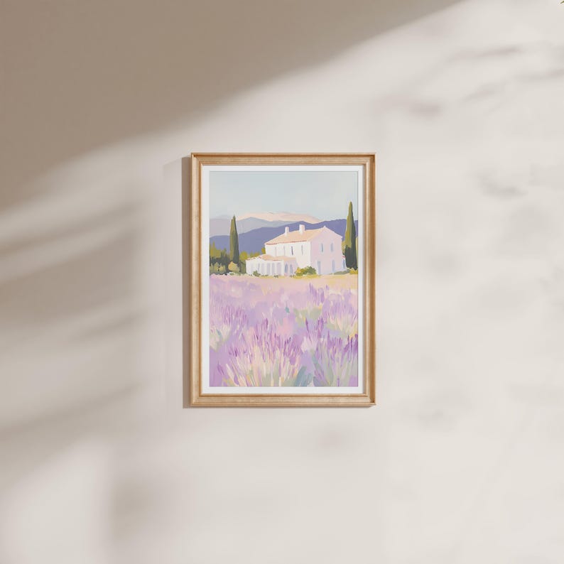 Framed artwork of a serene villa surrounded by lavender fields, hung on a softly lit wall. The painting features soft pastel tones with purple blooms, green cypress trees, and distant mountains under a pale sky.