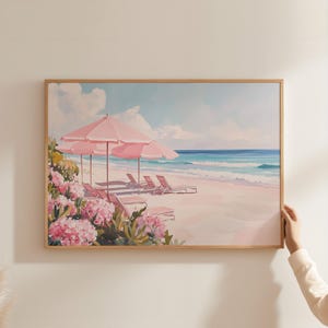 Coastal print featuring a tranquil pink beach scene being carefully hung on a light wall. A hand steadies the golden-framed artwork, which shows rows of lounge chairs, umbrellas, and tropical blooms beside a calm turquoise sea.
