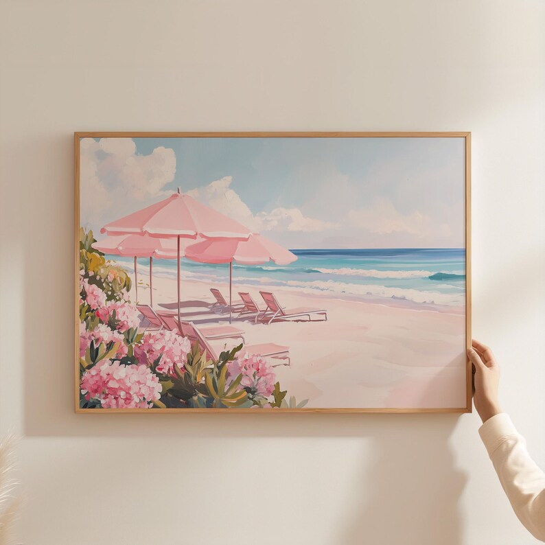 Coastal print featuring a tranquil pink beach scene being carefully hung on a light wall. A hand steadies the golden-framed artwork, which shows rows of lounge chairs, umbrellas, and tropical blooms beside a calm turquoise sea.