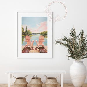 May include: A framed watercolor print depicts two pink Adirondack chairs on a wooden dock overlooking a lake. The sky is a blend of blue and pink, with trees lining the distant shore. The print is displayed above a white bench with three woven baskets.