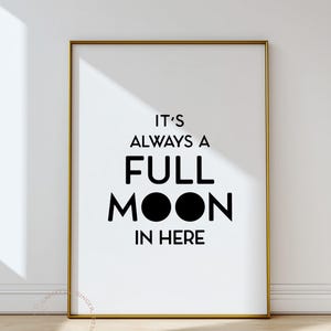 May include: A framed print with a gold border features the phrase "IT'S ALWAYS A FULL MOON IN HERE" in bold black letters on a white background. The design includes two black circles representing the moon.