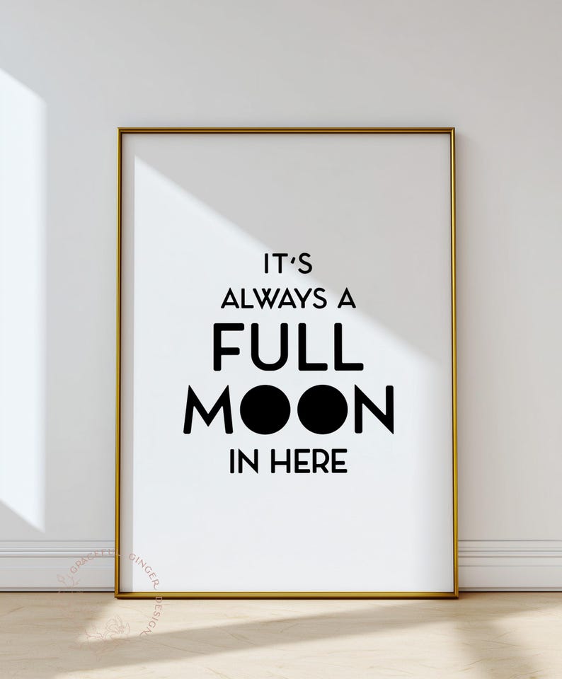 May include: A framed print with a gold border features the phrase "IT'S ALWAYS A FULL MOON IN HERE" in bold black letters on a white background. The design includes two black circles representing the moon.