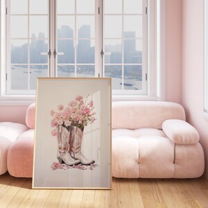 May include: A framed print of a still life featuring two white cowboy boots with pink flowers inside. The boots are on a white background with pink flower petals scattered around them.