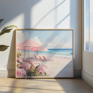 Framed beach artwork propped near a window where sunlight streams across the floor. The scene includes blush-toned umbrellas, beach chairs, and blooming flowers along a peaceful seashore with waves breaking gently in the distance.