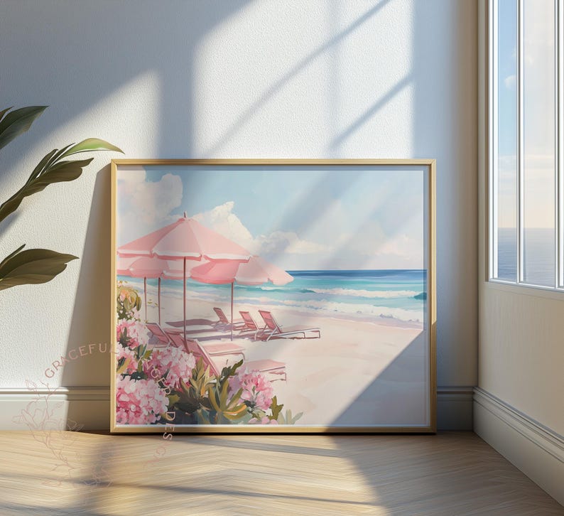 Framed beach artwork propped near a window where sunlight streams across the floor. The scene includes blush-toned umbrellas, beach chairs, and blooming flowers along a peaceful seashore with waves breaking gently in the distance.