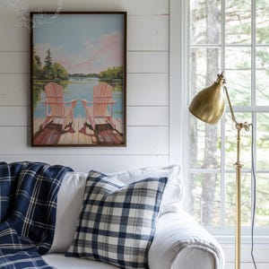 May include: A cozy living room scene featuring a white sofa adorned with a blue and white plaid pillow and a matching throw blanket. A framed painting of Adirondack chairs on a dock hangs above the sofa. A brass floor lamp stands beside a window.