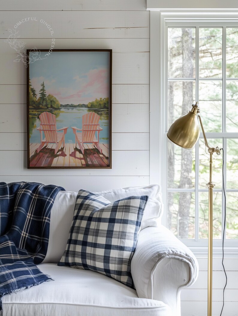 May include: A cozy living room scene featuring a white sofa adorned with a blue and white plaid pillow and a matching throw blanket. A framed painting of Adirondack chairs on a dock hangs above the sofa. A brass floor lamp stands beside a window.