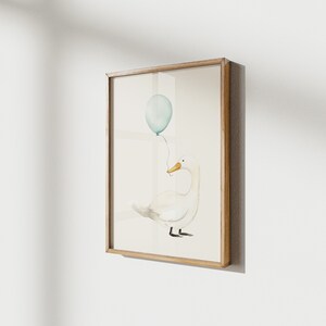 May include: Watercolor illustration of a white duck holding a light blue balloon. The artwork is framed in a light brown wooden frame and mounted on a white wall. The duck has a yellow beak and black feet.