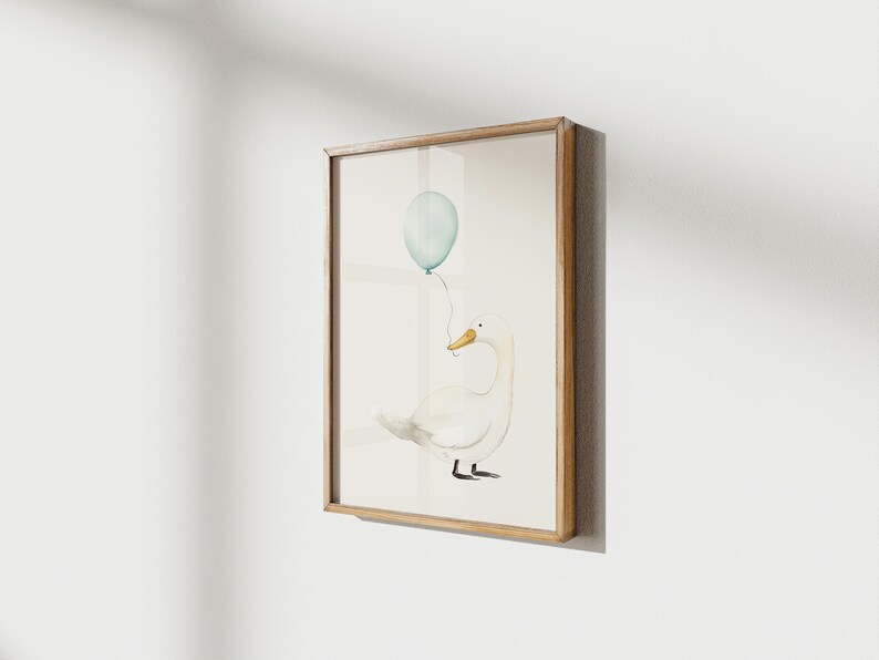 May include: Watercolor illustration of a white duck holding a light blue balloon. The artwork is framed in a light brown wooden frame and mounted on a white wall. The duck has a yellow beak and black feet.