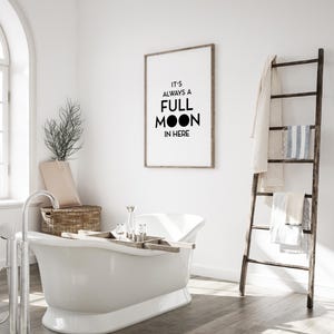 May include: A white clawfoot bathtub with a wooden tray holding bath accessories. A framed print on the wall reads "IT'S ALWAYS A FULL MOON IN HERE." A wooden ladder holds towels. The bathroom has a neutral color palette.