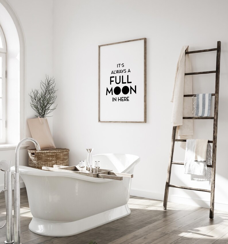 May include: A white clawfoot bathtub with a wooden tray holding bath accessories. A framed print on the wall reads "IT'S ALWAYS A FULL MOON IN HERE." A wooden ladder holds towels. The bathroom has a neutral color palette.