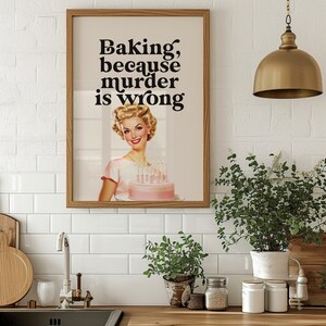 Peut inclure: Une impression encadr&eacute;e avec une illustration r&eacute;tro d'une femme tenant un g&acirc;teau rose avec des bougies. Le texte sur l'impression dit "Baking, because murder is wrong."