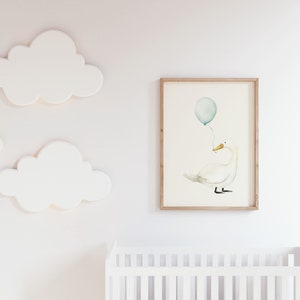 May include: A framed watercolor print of a white swan holding a teal balloon. The artwork is displayed on a white wall, alongside three white cloud-shaped wall decorations. A white crib is visible in the foreground.