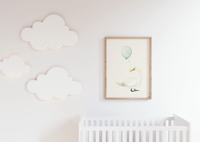May include: A framed watercolor print of a white swan holding a teal balloon. The artwork is displayed on a white wall, alongside three white cloud-shaped wall decorations. A white crib is visible in the foreground.
