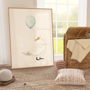 May include: Watercolor print of a white duck holding a teal balloon. The artwork is framed in a light wood frame. The scene is set in a nursery with a wicker basket, a stuffed animal, and a decorative pillow.