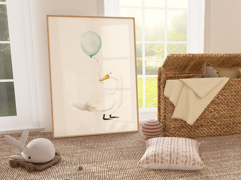 May include: Watercolor print of a white duck holding a teal balloon. The artwork is framed in a light wood frame. The scene is set in a nursery with a wicker basket, a stuffed animal, and a decorative pillow.