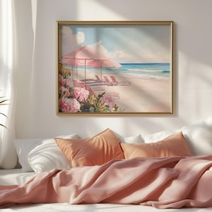 Beach-themed painting hung above a cozy bed styled with soft coral pillows and blankets. The art captures a pastel paradise with pink umbrellas and flowers beside an ocean shoreline, bathed in warm morning light.