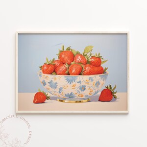 Op de afbeelding: Een witte kom met een blauw bloemenpatroon gevuld met rode aardbeien. De kom staat op een lichtblauw oppervlak met een enkele aardbei ervoor.