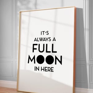 May include: A framed art print with the text "IT'S ALWAYS A FULL MOON IN HERE" in black. The print has a white background and a gold frame with an orange edge. It leans against a wall with a shadow.