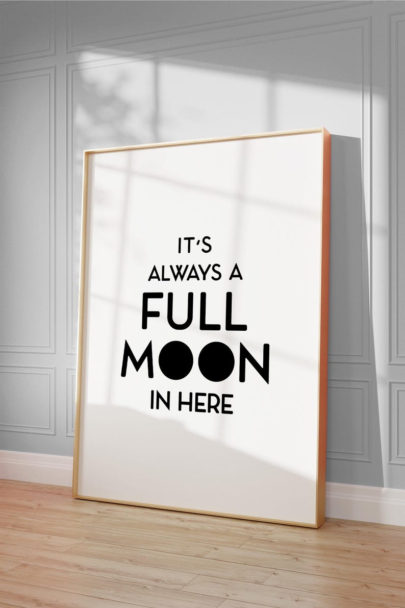 May include: A framed art print with the text "IT'S ALWAYS A FULL MOON IN HERE" in black. The print has a white background and a gold frame with an orange edge. It leans against a wall with a shadow.