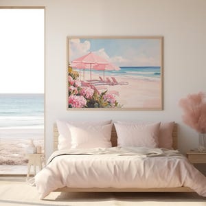 Coastal-themed beach print displayed above a soft pink bed in a light-filled bedroom. The artwork features pink umbrellas and flowers beside tranquil turquoise waters, echoing the serene ocean view just outside the open door.