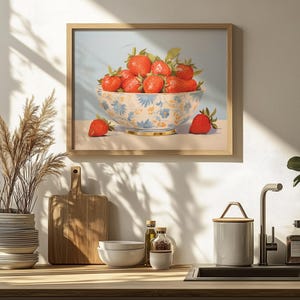 A framed painting of a bowl filled with strawberries, featuring a delicate floral pattern on the bowl. The artwork hangs on a kitchen wall, with soft natural light highlighting the vibrant reds of the strawberries against the pale blue background.