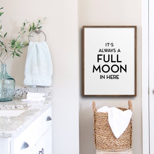 May include: A bathroom scene with a white vanity, a framed sign that reads "IT'S ALWAYS A FULL MOON IN HERE", and a woven basket. A light blue towel hangs on a silver ring, and a glass vase holds green branches. A white door is on the right.