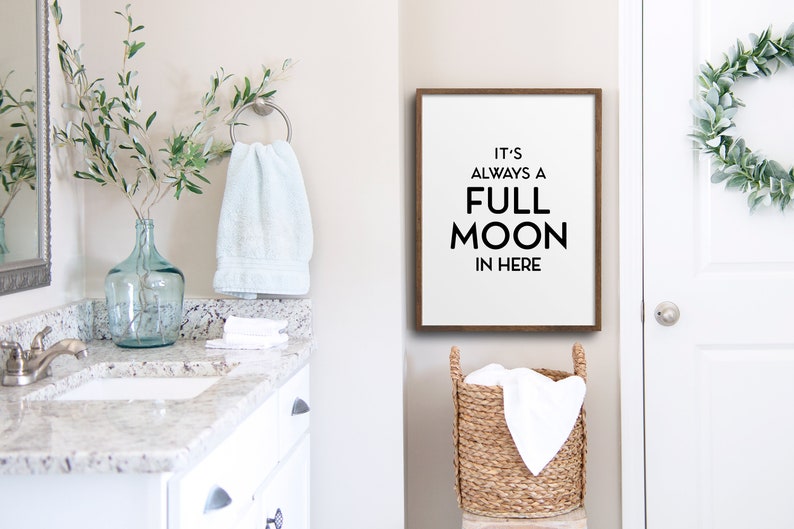 May include: A bathroom scene with a white vanity, a framed sign that reads "IT'S ALWAYS A FULL MOON IN HERE", and a woven basket. A light blue towel hangs on a silver ring, and a glass vase holds green branches. A white door is on the right.