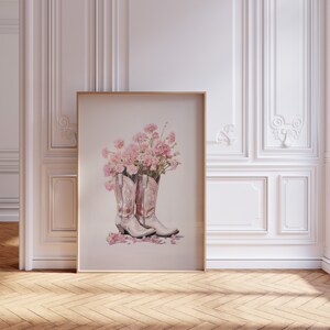 May include: A framed print of a watercolor painting of a pair of white cowboy boots with pink carnations inside. The boots are on a white background with pink petals scattered around them.