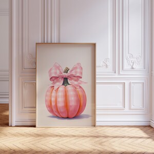 Puede incluir: Una impresi&oacute;n enmarcada con una calabaza rosa con un lazo de cuadros. La calabaza tiene un degradado de tonos rosas y naranjas, con un tallo verde. El lazo es de un patr&oacute;n de cuadros rosa claro y blanco. La obra de arte se exhibe en una habitaci&oacute;n con paredes blancas y suelo de madera.