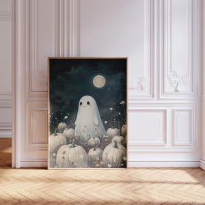May include: A framed art print featuring a friendly ghost in a field of pumpkins under a full moon. The artwork has a whimsical, cartoonish style with a dark blue night sky and a light color palette. The print is in a wooden frame.