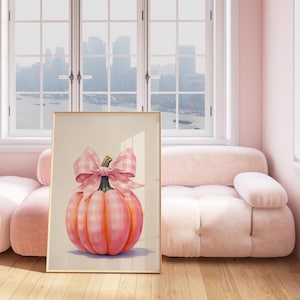 Puede incluir: Una impresi&oacute;n art&iacute;stica enmarcada que presenta una pintura a la acuarela de una calabaza rosa con un lazo de cuadros. La calabaza tiene un patr&oacute;n a cuadros y est&aacute; colocada sobre un fondo neutro. La obra de arte se exhibe en una habitaci&oacute;n luminosa con un sof&aacute; rosa.