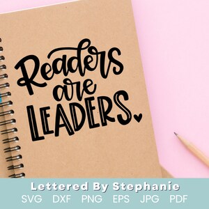May include: A brown notebook with a spiral binding and the text "Readers are Leaders" written in black cursive font. The notebook is on a pink surface.