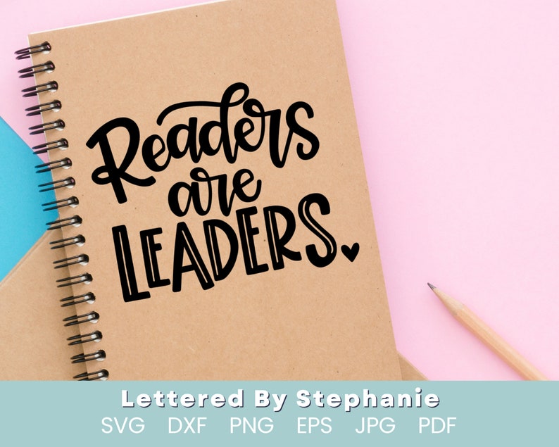 May include: A brown notebook with a spiral binding and the text "Readers are Leaders" written in black cursive font. The notebook is on a pink surface.