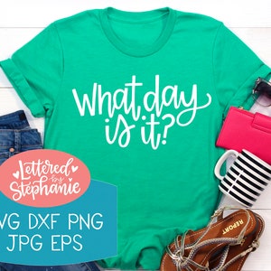 May include: A green t-shirt with the text "What day is it?" in white lettering. The shirt is laying on a white wooden surface with other items, including a pair of jeans, a pink wallet, sunglasses, a coffee mug, and sandals. The text "Lettered by Stephanie" is in a pink circle with white lettering and hearts. Below the text is a blue rectangle with the text "SVG DXF PNG JPG EPS" in white lettering.