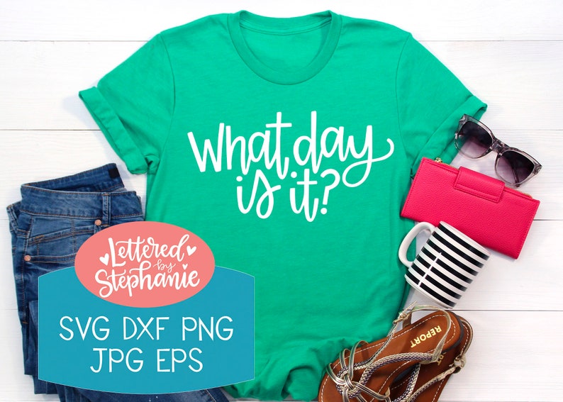 May include: A green t-shirt with the text "What day is it?" in white lettering. The shirt is laying on a white wooden surface with other items, including a pair of jeans, a pink wallet, sunglasses, a coffee mug, and sandals. The text "Lettered by Stephanie" is in a pink circle with white lettering and hearts. Below the text is a blue rectangle with the text "SVG DXF PNG JPG EPS" in white lettering.