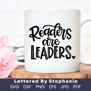 May include: White ceramic mug with a black design that says "Readers are Leaders" with a small heart at the end. The mug is sitting on a wooden surface.