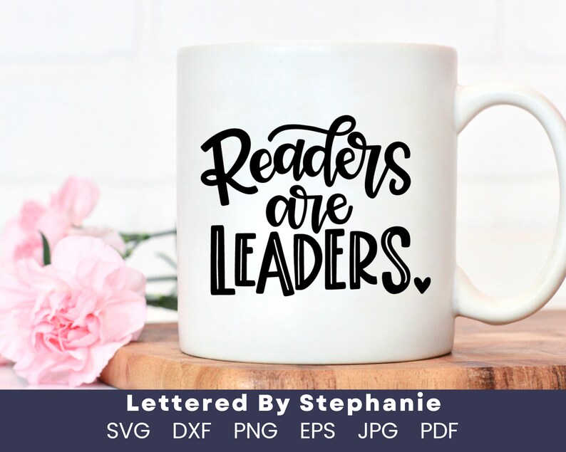 May include: White ceramic mug with a black design that says "Readers are Leaders" with a small heart at the end. The mug is sitting on a wooden surface.